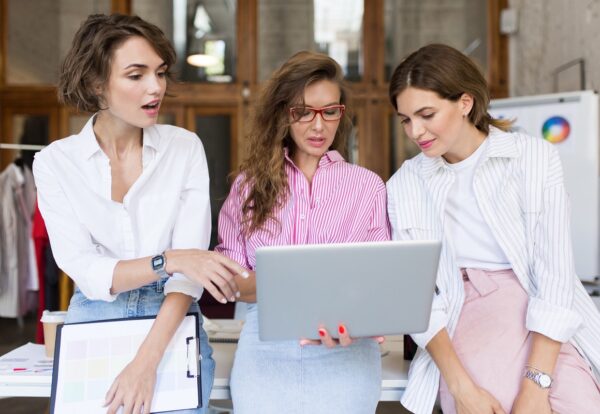 Group of young stylish women amazedly working on laptop together in modern cozy office. Thoughtful girls spending time at work